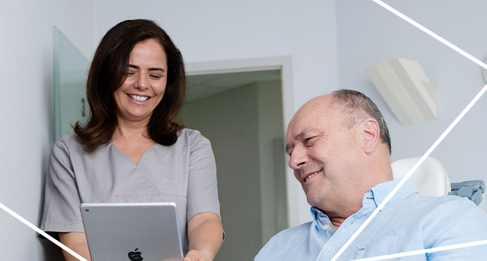 ZFA hält ein Tablet in der Hand und schaut gemeinsam mit einem auf dem Behandlungsstuhl sitzenden Patienten darauf
